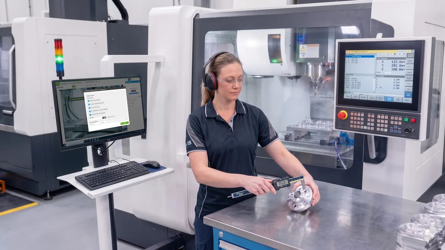 Opératrice dans une cellule d’usinage CNC avec un écran VKS et un panneau de commande machine à côté d’elle.