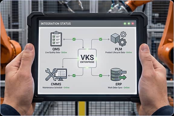 Mains tenant une tablette affichant l’écran radar d’intégrations VKS avec des nœuds ERP, PLM et QMS, près de bras robotisés